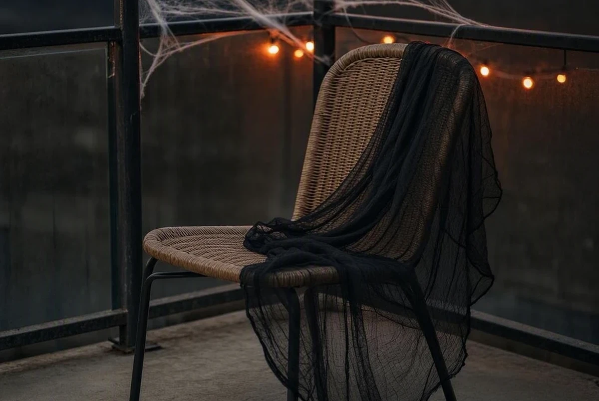 Black cheesecloth mesh draped loosely over the arm of an outdoor balcony chair, faux cobwebs stretched above between two railing posts, warm string lights glowing softly in the background